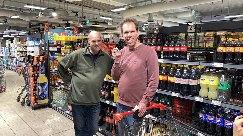 "Thilo steht mit einer anderen Person im REWE vor dem Getränkeregal."