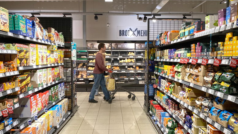 "Thilo geht im Rewe mit dem Einkaufswagen zwischen zwei Gängen entlang"