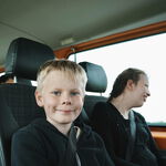 Ein lachender Junge sitzt in einem orangen Bus. Neben ihm eine Mitfahrerin, die lächelnd aus dem Fenster guckt. Hinter ihnen sitzen noch zwei Kinder